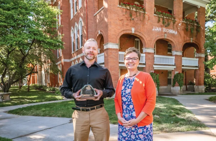 Two people posing for a picture outside of a brick building holding an award.